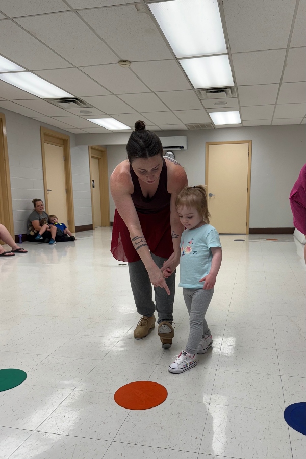 An adult guides a toddler during a movement activity on a marked floor in a studio.