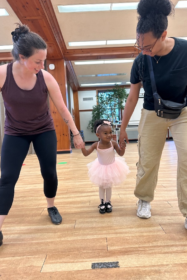 A young toddler in a pink tutu holds hands with two adults in a dance studio, smiling and taking steps together on a wooden floor.