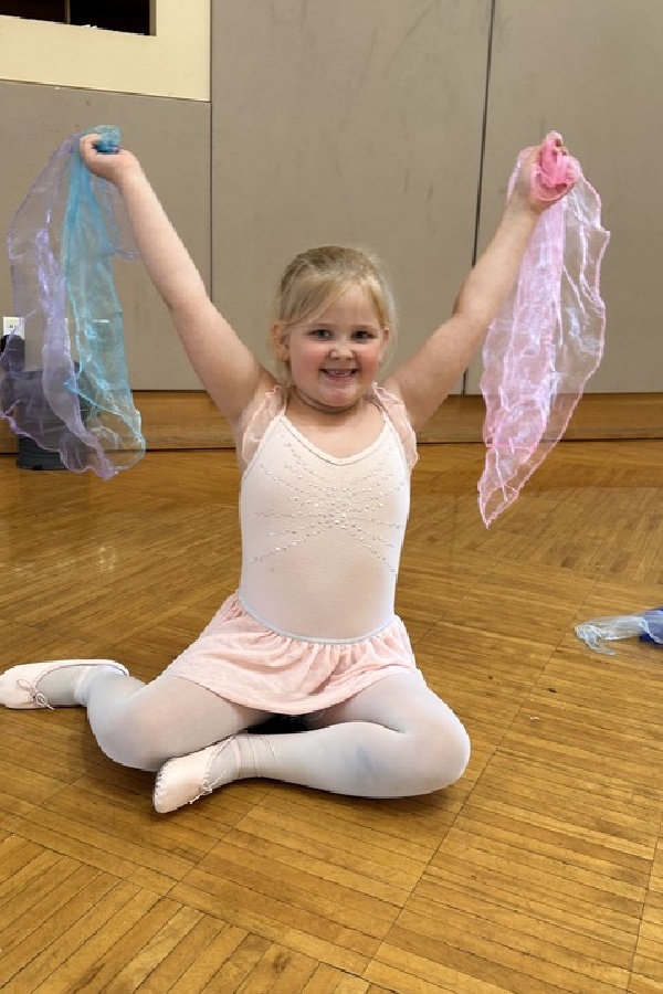 Young girl in a pink ballet outfit sitting on a wooden floor with legs crossed, smiling and holding up two colorful scarves, one blue and one pink.
