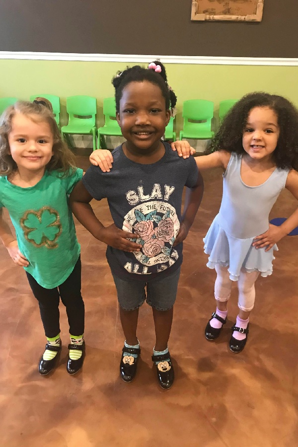 Three young girls stand smiling on a brown floor, wearing tap shoes. They pose confidently with green chairs behind them.