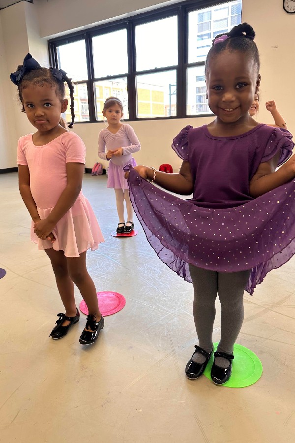 Three young girls in ballet attire stand on colored floor spots in a dance studio, smiling and posing for the camera.