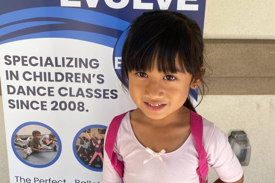 Young girl in a pink dance outfit and backpack smiles in front of a dance school banner promoting children’s dance classes.