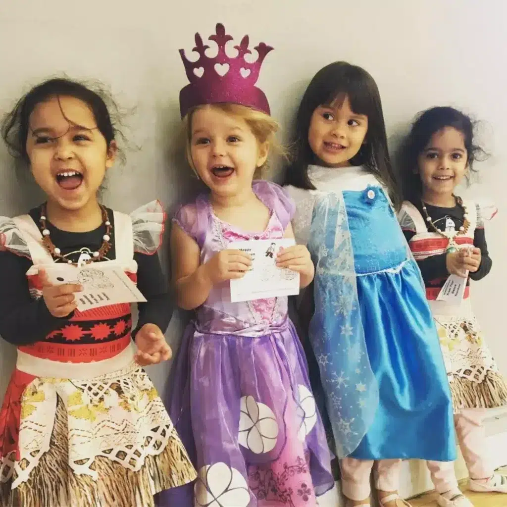 Four young girls dressed in colorful costumes, smiling and posing together. One girl is wearing a purple princess dress with a crown, another in a blue Elsa dress, one in a Moana-inspired outfit, and another in a red and white dress with a skirt, all holding coloring cards.
